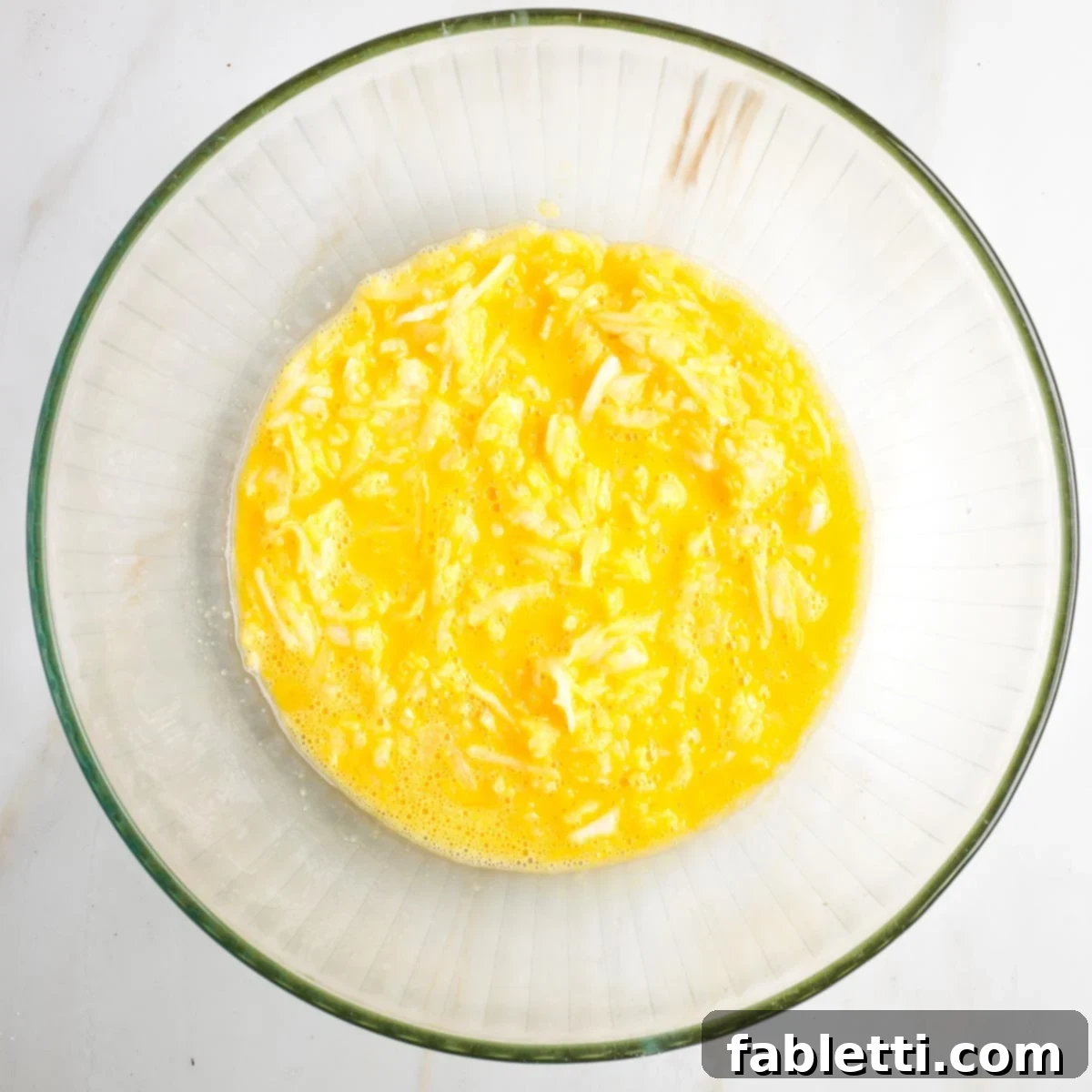 Grandma's Golden Potato Latkes 5 Frothy whisked eggs mixed with finely shredded onions and their juices in a clear glass mixing bowl.