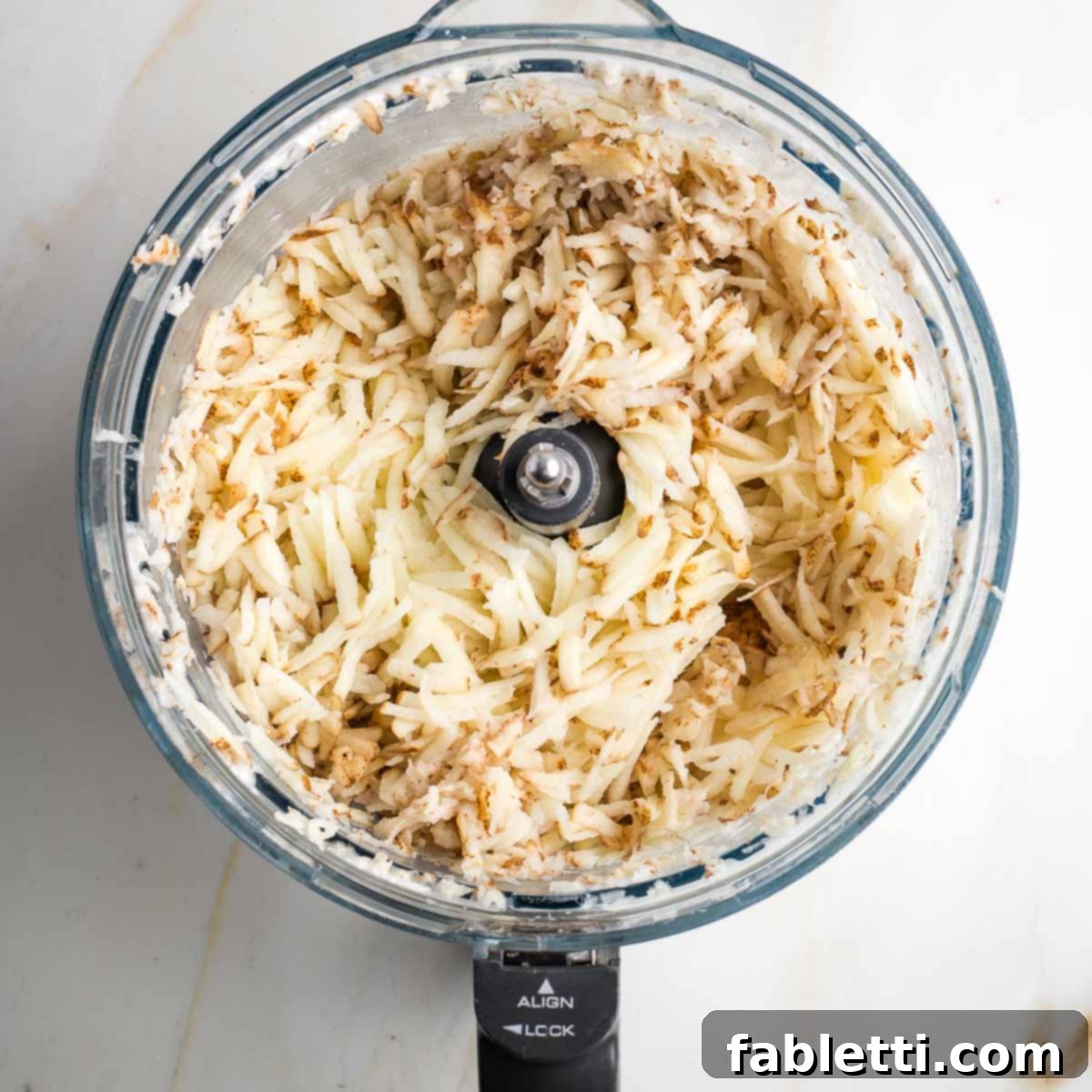 Grandma's Golden Potato Latkes 7 Freshly shredded russet potatoes in the clean bowl of a food processor, ready to be mixed.