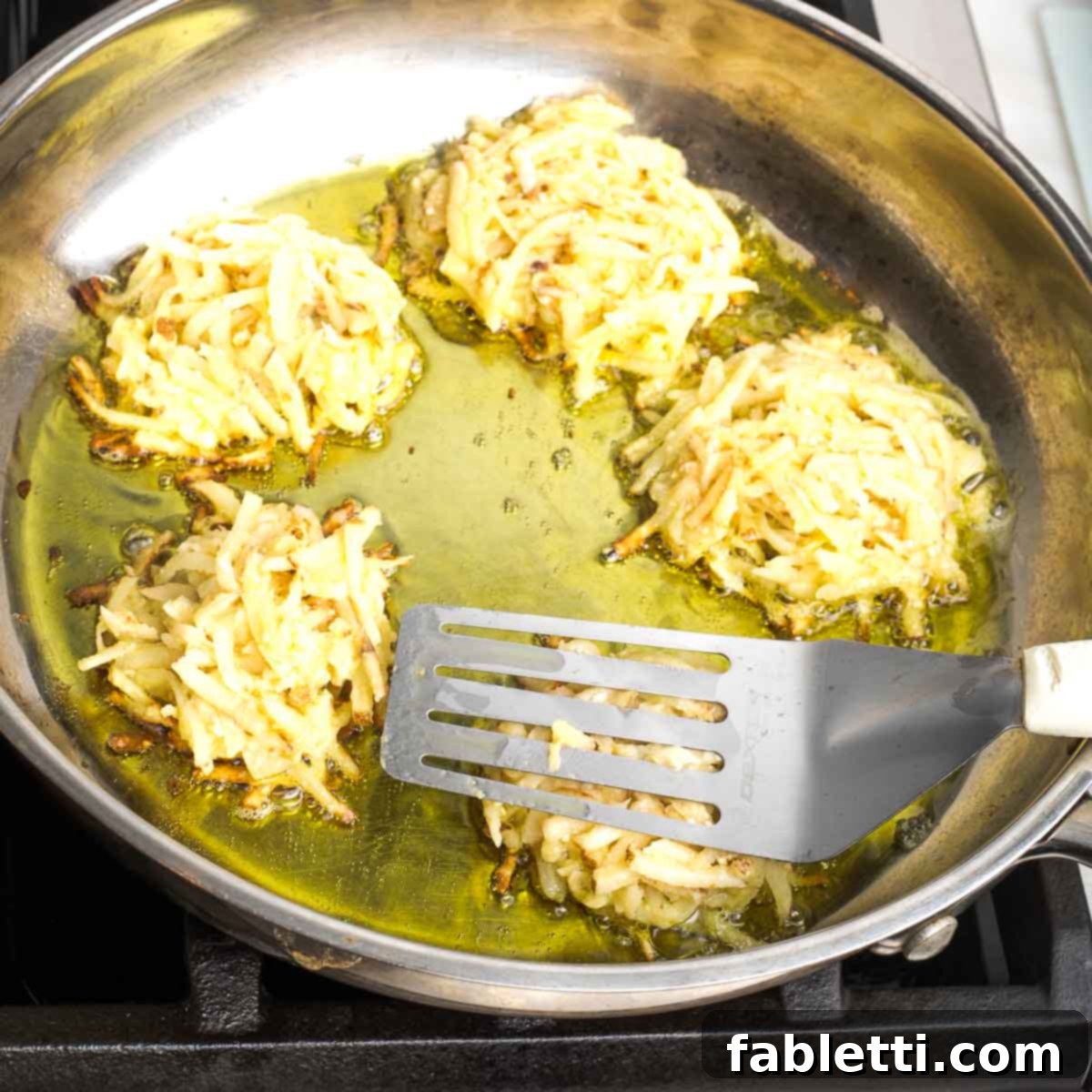 Grandma's Golden Potato Latkes 10 A spatula pressing down on a potato latke in a large skillet, surrounded by other frying latkes in hot oil.