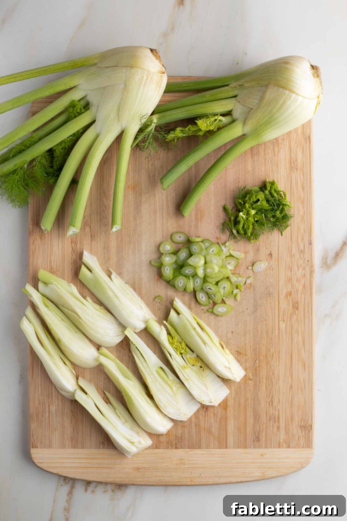 Neatly arranged fennel wedges lying on their sides, accompanied by finely sliced stalks and a small pile of fresh fronds, ready for cooking and garnishing.
