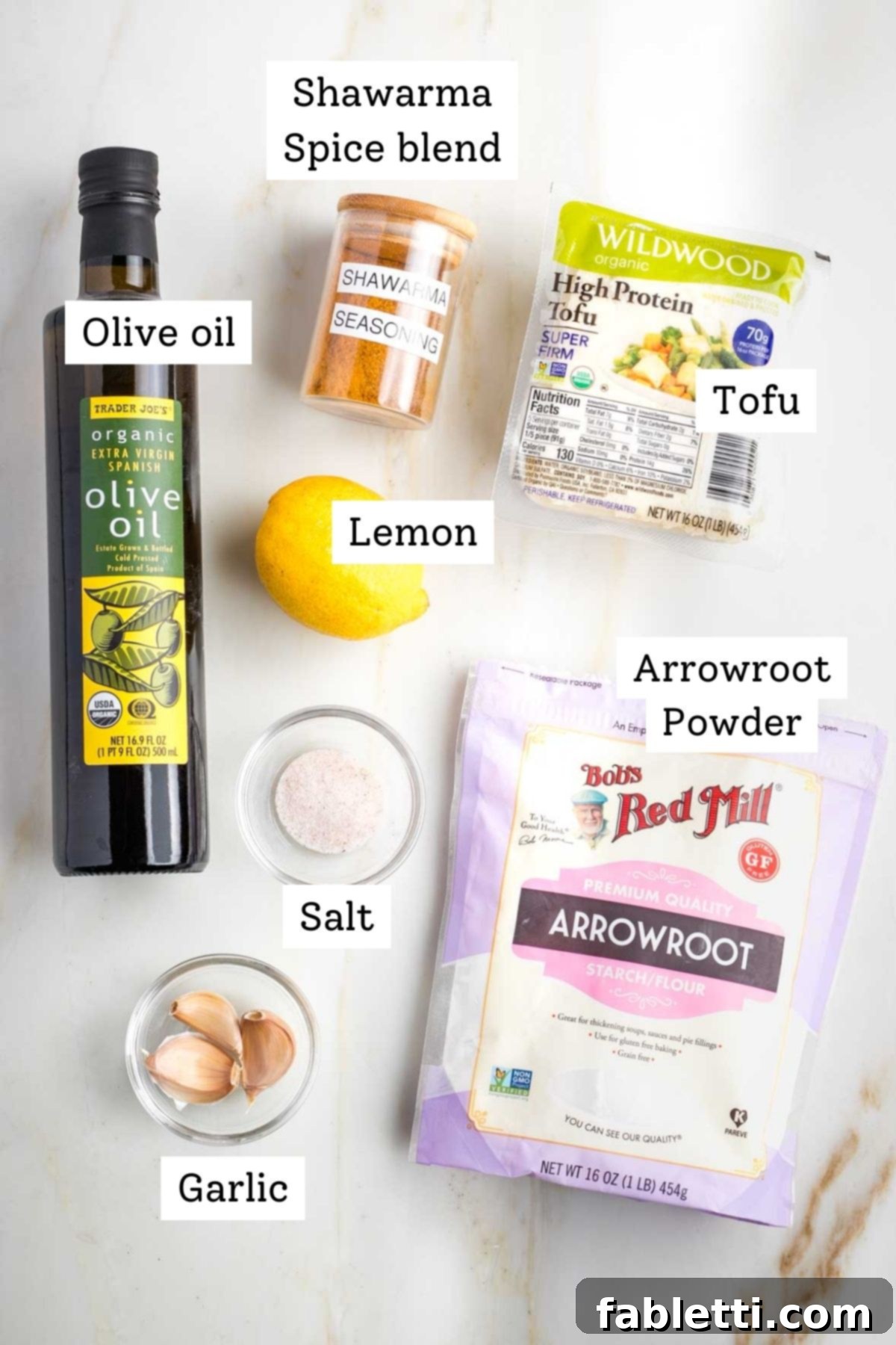 Flavorful Vegan Tofu Shawarma 4 Labeled ingredients for tofu shawarma: a bottle of olive oil, a bowl of shawarma spice blend, a block of tofu, a container of arrowroot powder, garlic cloves, a pinch of salt, and a fresh lemon.