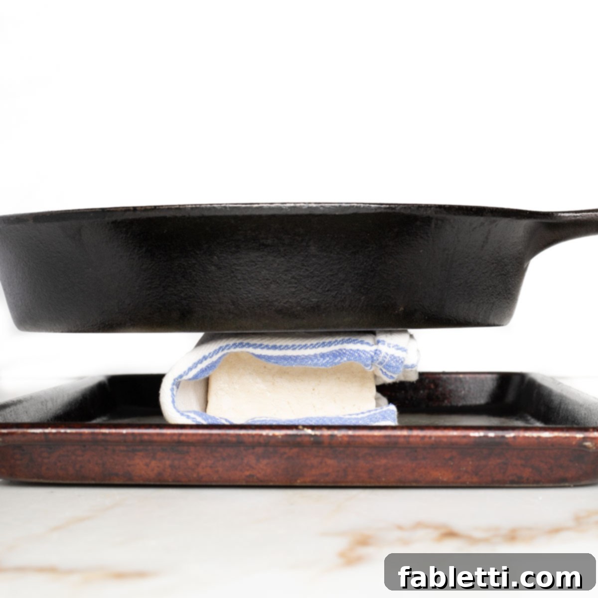 Flavorful Vegan Tofu Shawarma 5 A block of tofu, carefully wrapped in a clean kitchen towel, with a heavy cast iron skillet placed on top to press out excess water.
