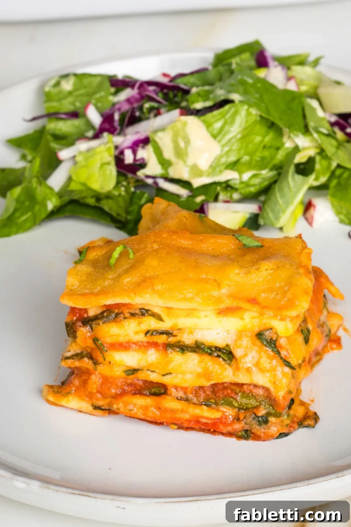 Lush Layers: Gluten-Free Vegan Garden Lasagna 16 Layers of lasagna noodles, bechamel, red sauce, spinach and zucchini shown from the side with a green salad in the background.