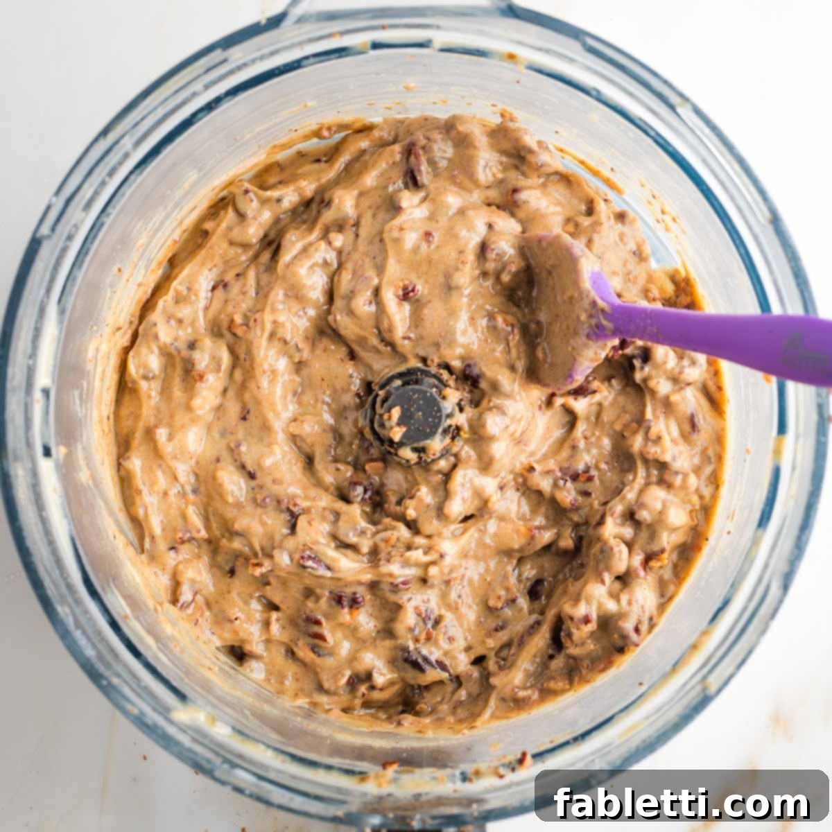 Naturally Sweetened Vegan Pecan Pie Bars 16 Golden colored caramel spread with chopped pecans in the bowl of a food processor with silicon spatula stirring.