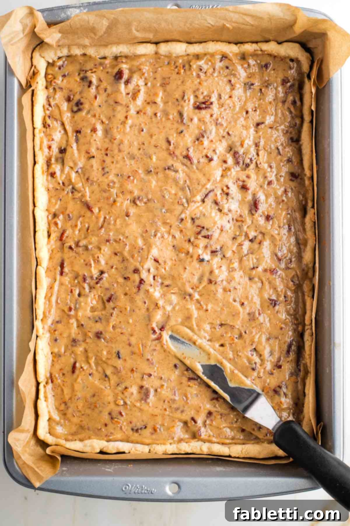 Naturally Sweetened Vegan Pecan Pie Bars 18 Offset spatula spreading caramel pecan spread onto a pie crust in a rectangular baking pan.
