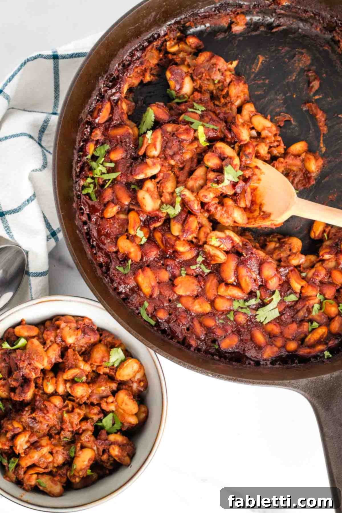 Effortless Skillet Plant-Based BBQ Beans 2 A rustic cast iron skillet brimming with vibrant BBQ baked beans, generously garnished with fresh herbs. A wooden spoon carefully scoops a portion into a small white bowl, ready for serving.