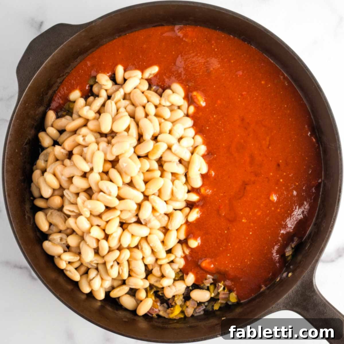 Effortless Skillet Plant-Based BBQ Beans 11 The cast iron skillet, previously holding sautéed vegetables, is now generously topped with rinsed canned beans and a rich, deep red BBQ sauce, ready for simmering and baking.