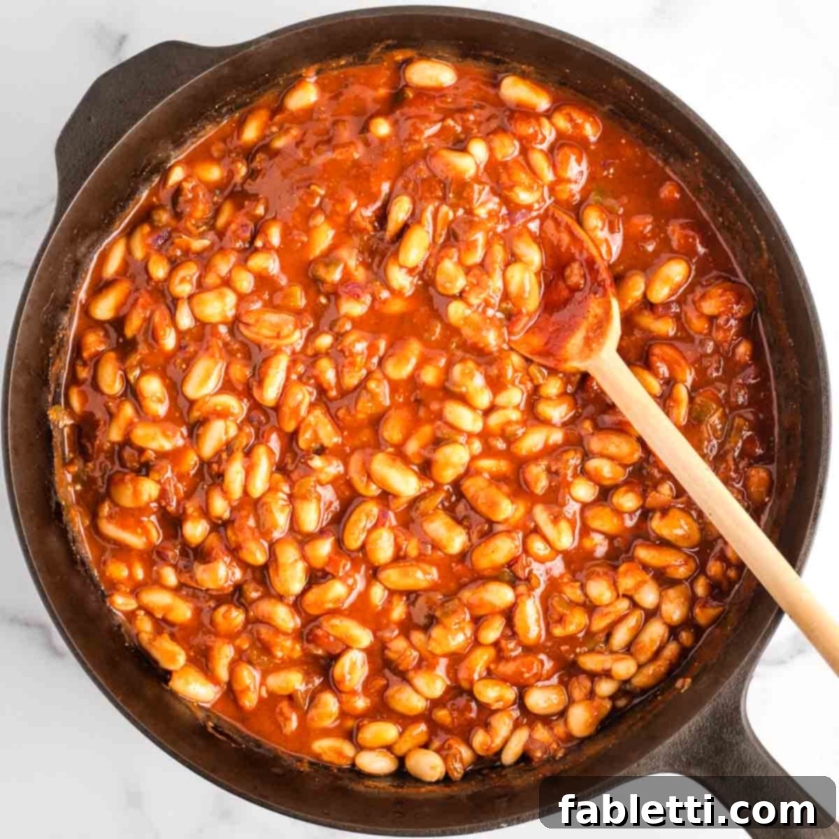 Effortless Skillet Plant-Based BBQ Beans 12 A wooden spoon stirs the cannellini beans, gently coating them in the thick, lustrous tomato-based BBQ sauce, ensuring every bean is richly flavored.