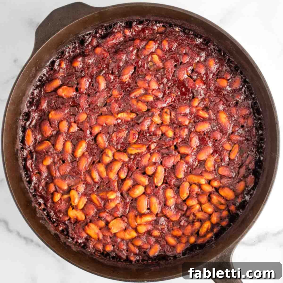 Effortless Skillet Plant-Based BBQ Beans 13 A close-up view of the cast iron skillet filled with bubbling baked beans, their deep red BBQ sauce glistening as it slowly thickens in the oven.