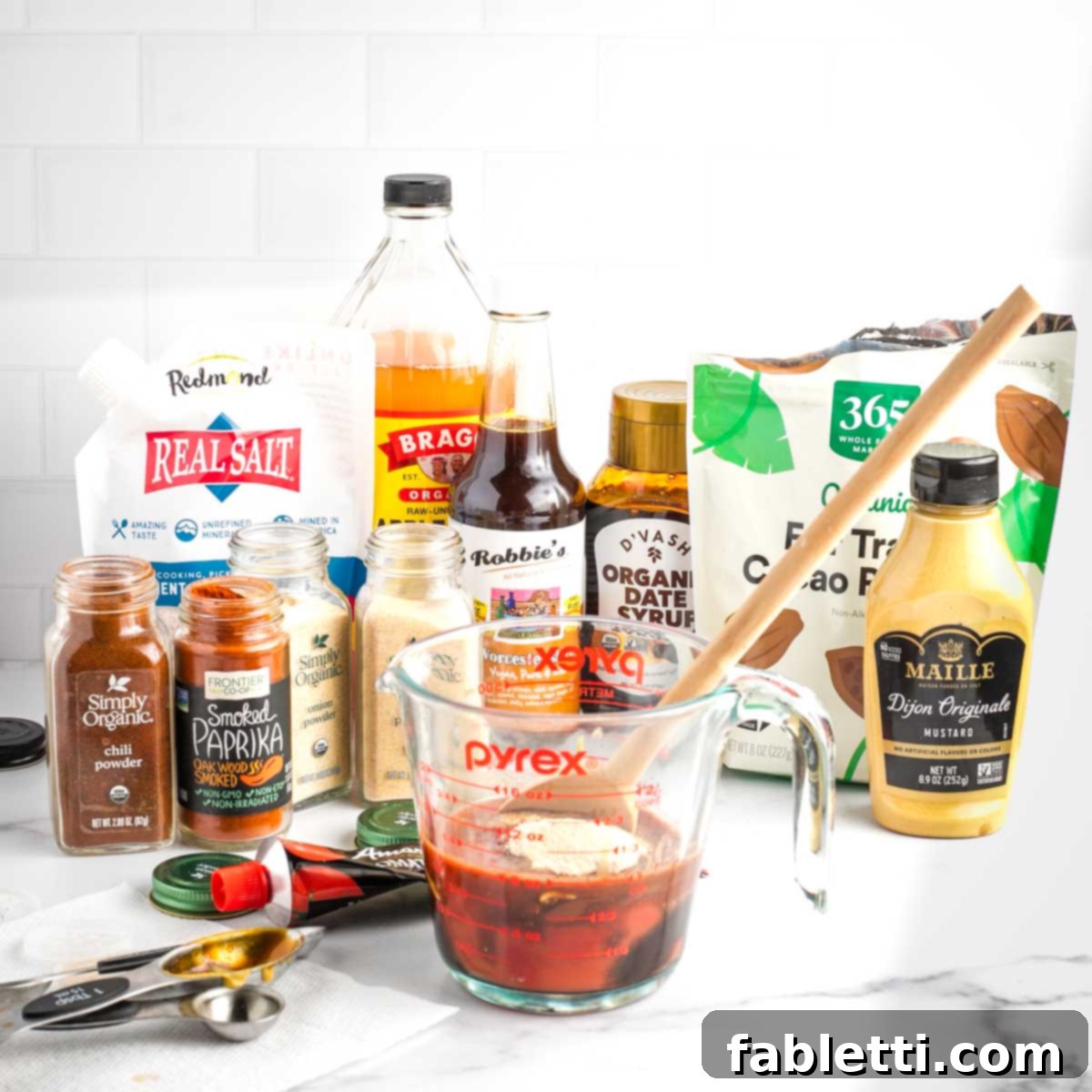 Effortless Skillet Plant-Based BBQ Beans 3 An array of bottles, jars, and spices neatly arranged around a Pyrex measuring cup, in which homemade BBQ sauce is being stirred to perfection.