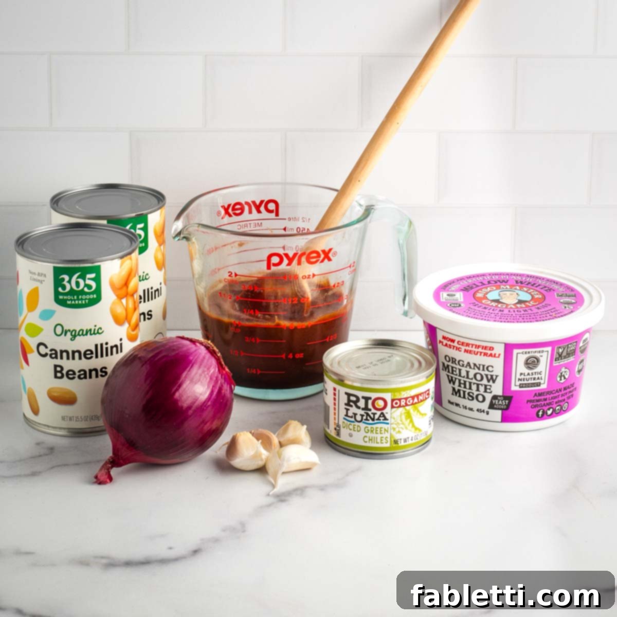 Effortless Skillet Plant-Based BBQ Beans 4 A visual display of essential ingredients: a measuring cup filled with rich BBQ sauce, alongside cans of cannellini beans, a vibrant red onion, whole garlic cloves, a can of green chiles, and a container of light miso paste, all neatly arranged for cooking.