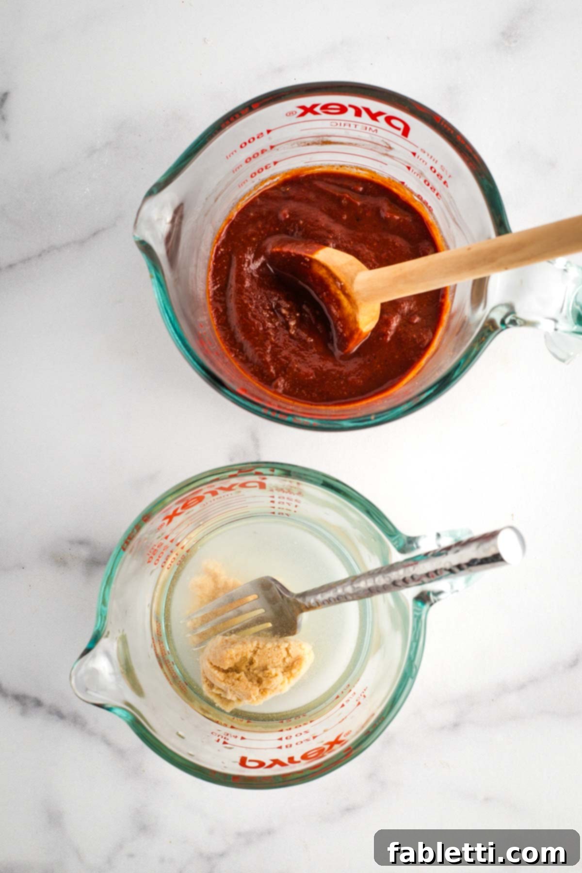 Effortless Skillet Plant-Based BBQ Beans 6 A measuring cup filled with rich BBQ sauce is shown next to a separate measuring cup containing miso paste dissolved in water, being stirred with a fork to achieve an even consistency.