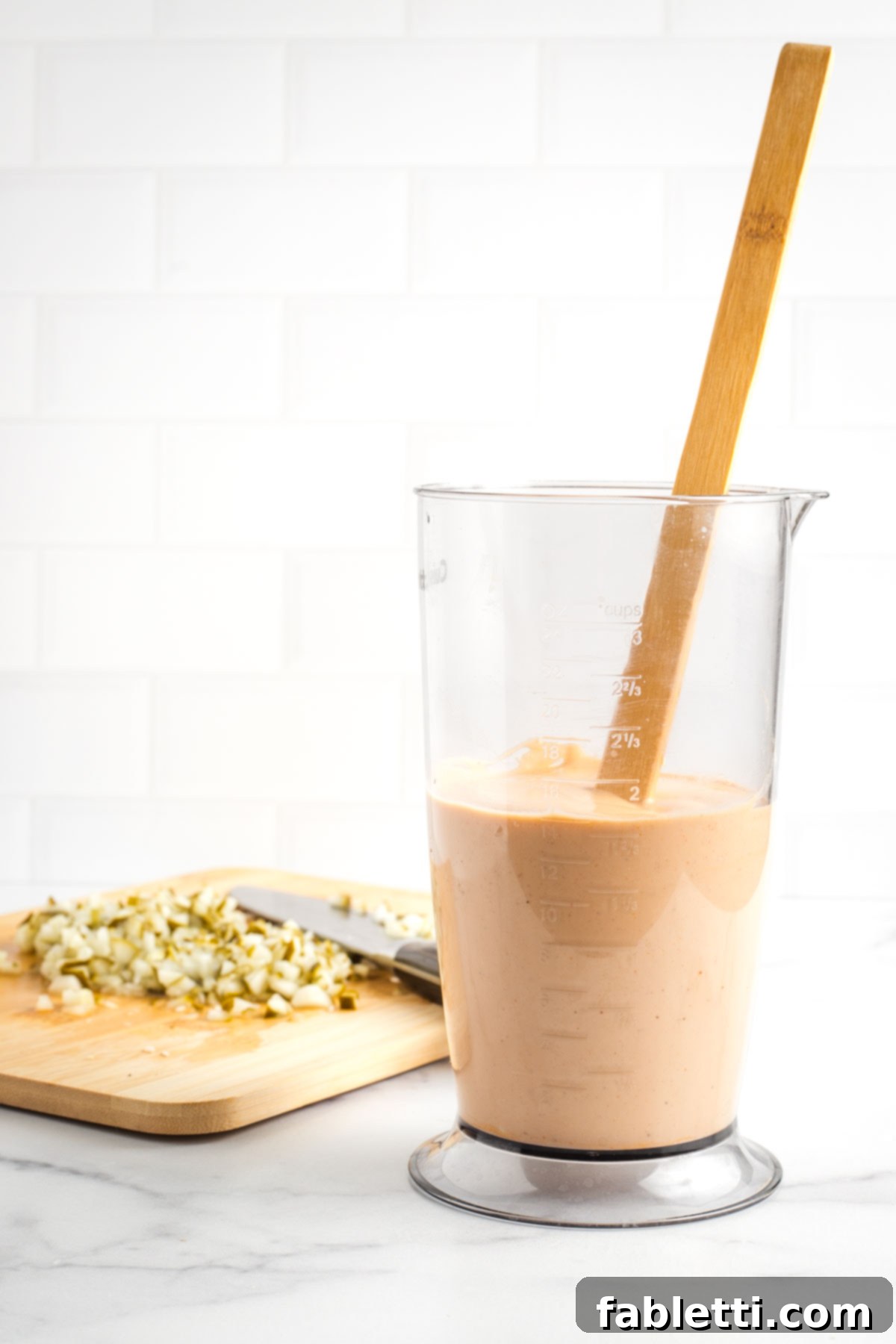 Creamy Tofu Vegan Big Mac Sauce 7 Orange burger sauce in a tall container with small diced pickles on a board in the background.