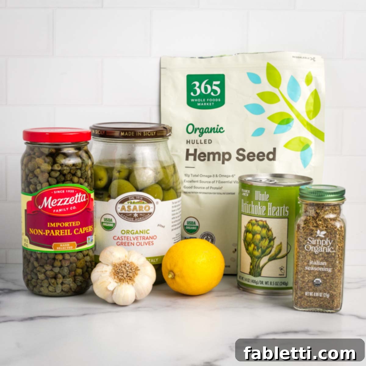 Ingredients for olive tapenade including green olives, capers, hemp seeds, garlic, lemon, artichoke hearts and italian seasoning. 