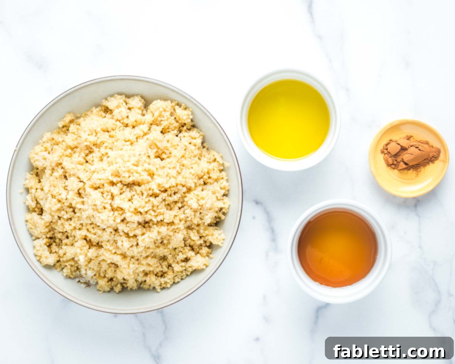 A bowl of cooked quinoa, alongside small dishes containing olive oil, maple syrup, and a generous scoop of ground cinnamon – ingredients for sweet crispy quinoa.