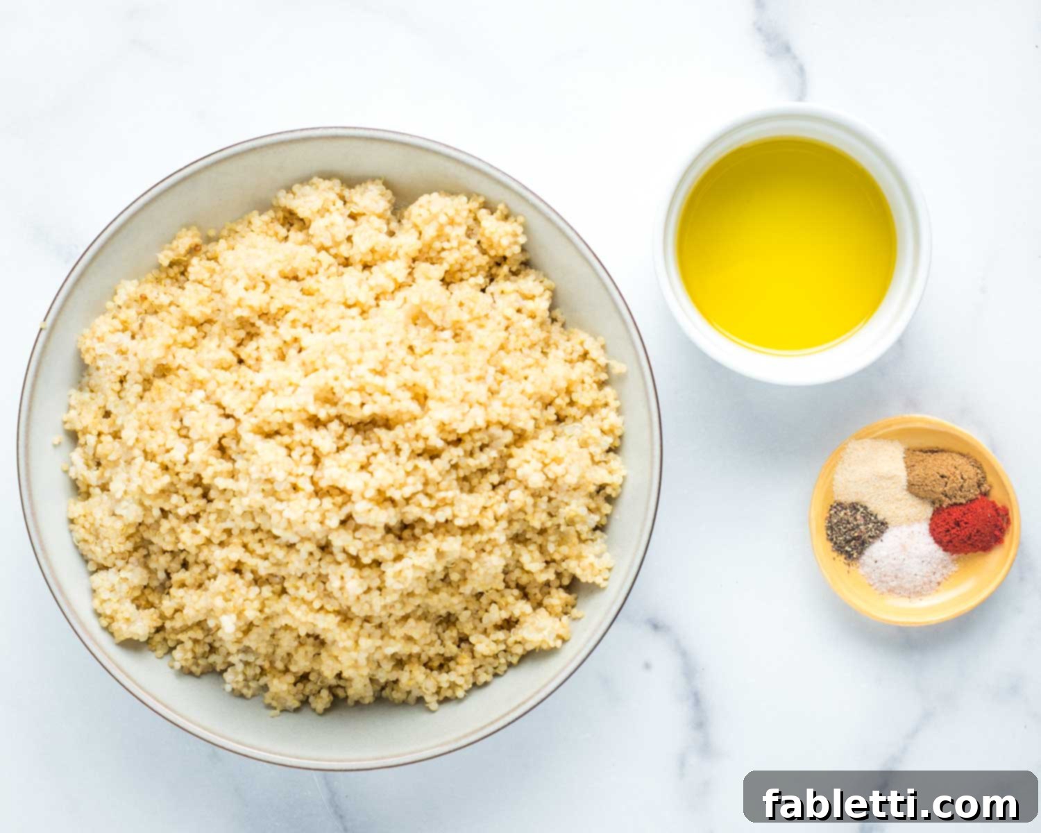 A bowl of cooked quinoa, a dish of olive oil, and an assortment of colorful savory spices, ready for mixing.