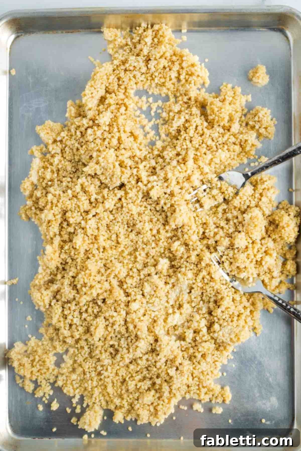 Two forks expertly spreading cooked quinoa out onto a large, rimmed baking sheet, ensuring an even layer.