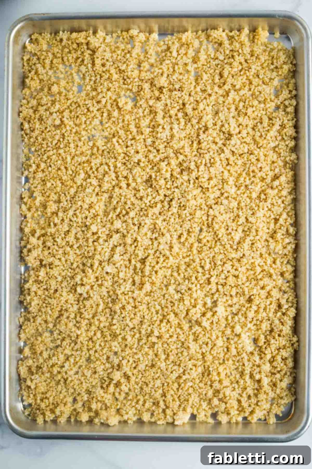 A large, rimmed baking sheet covered uniformly with cooked quinoa, ready for seasoning.
