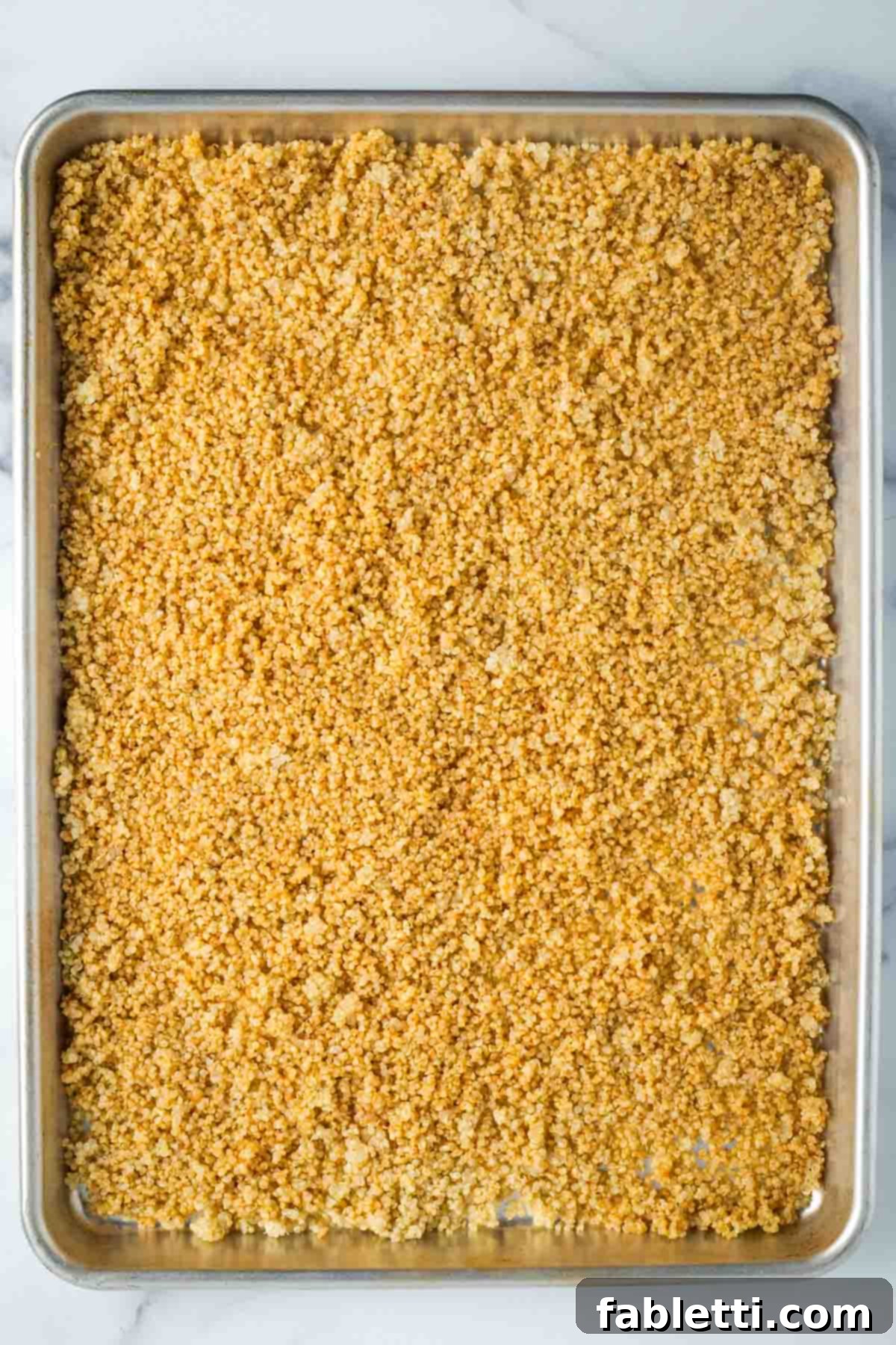 Cooked quinoa spread onto a large, rimmed baking sheet, now drizzled with olive oil and spices, awaiting its first bake.