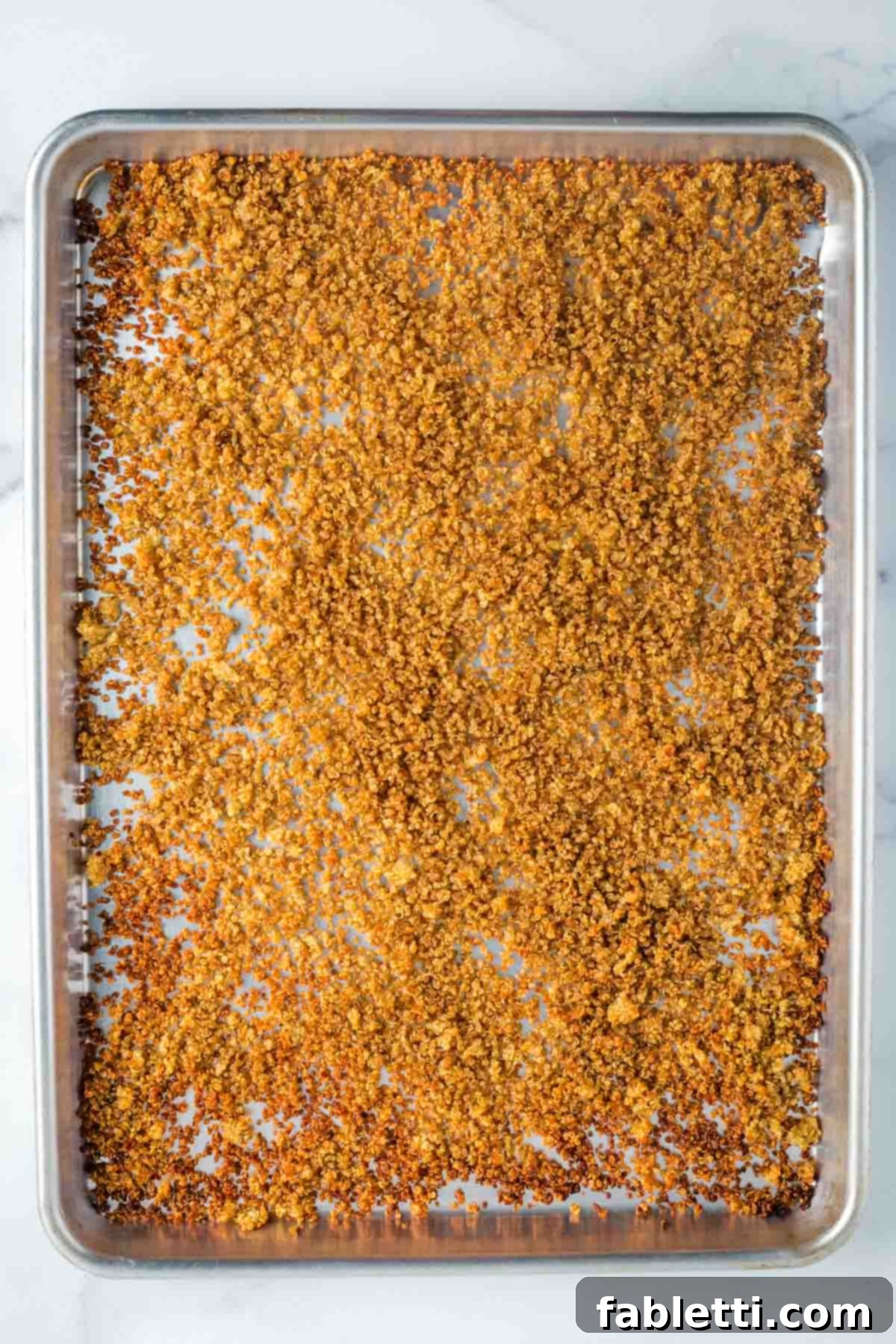 Golden, partially crispy quinoa spread onto a rimmed baking sheet, having been stirred and redistributed during the baking process.