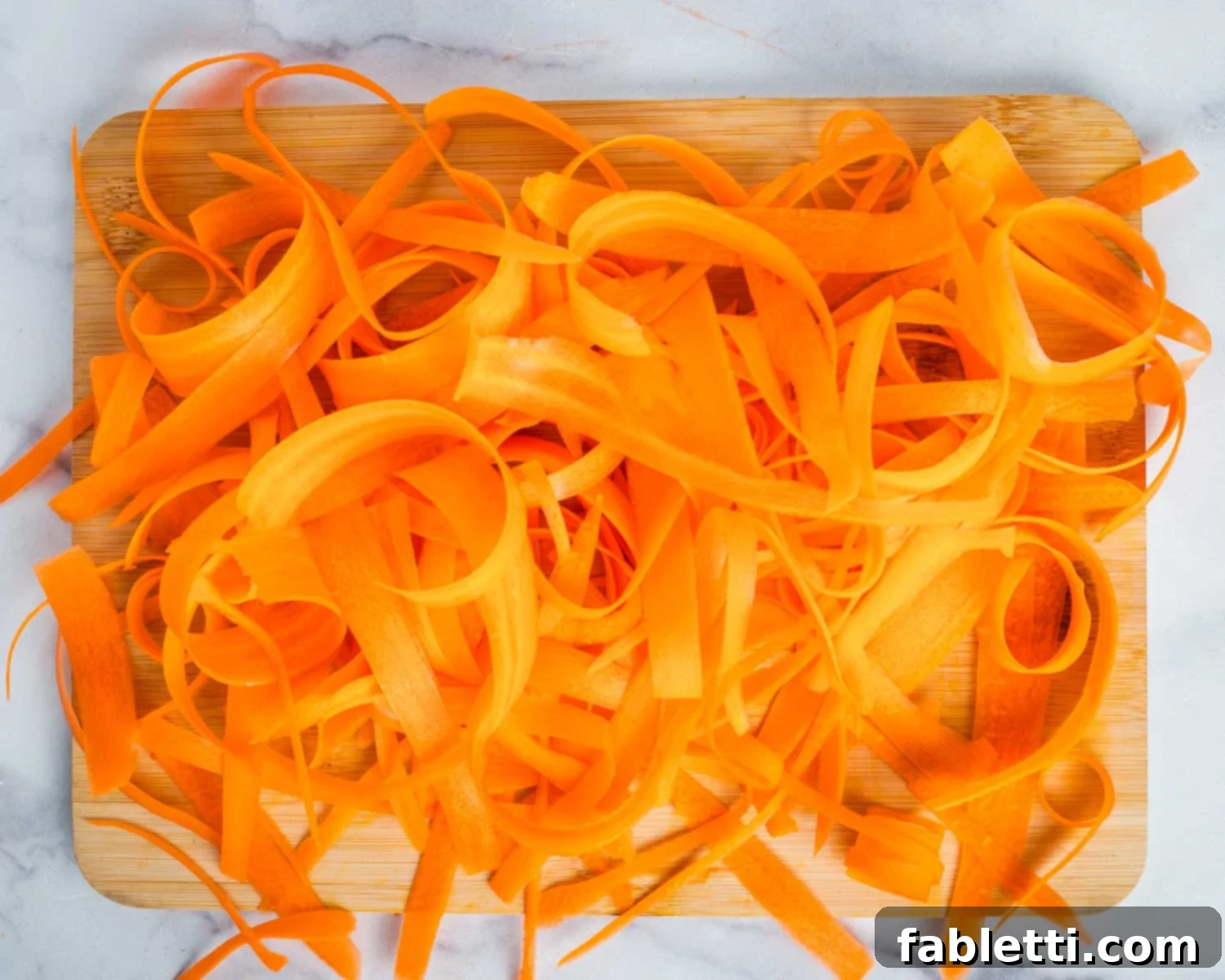Sweet Heat Shaved Carrot Salad 5 An inviting pile of long, thinly sliced carrot ribbons neatly stacked on a wooden cutting board, showcasing their vibrant color and delicate texture.