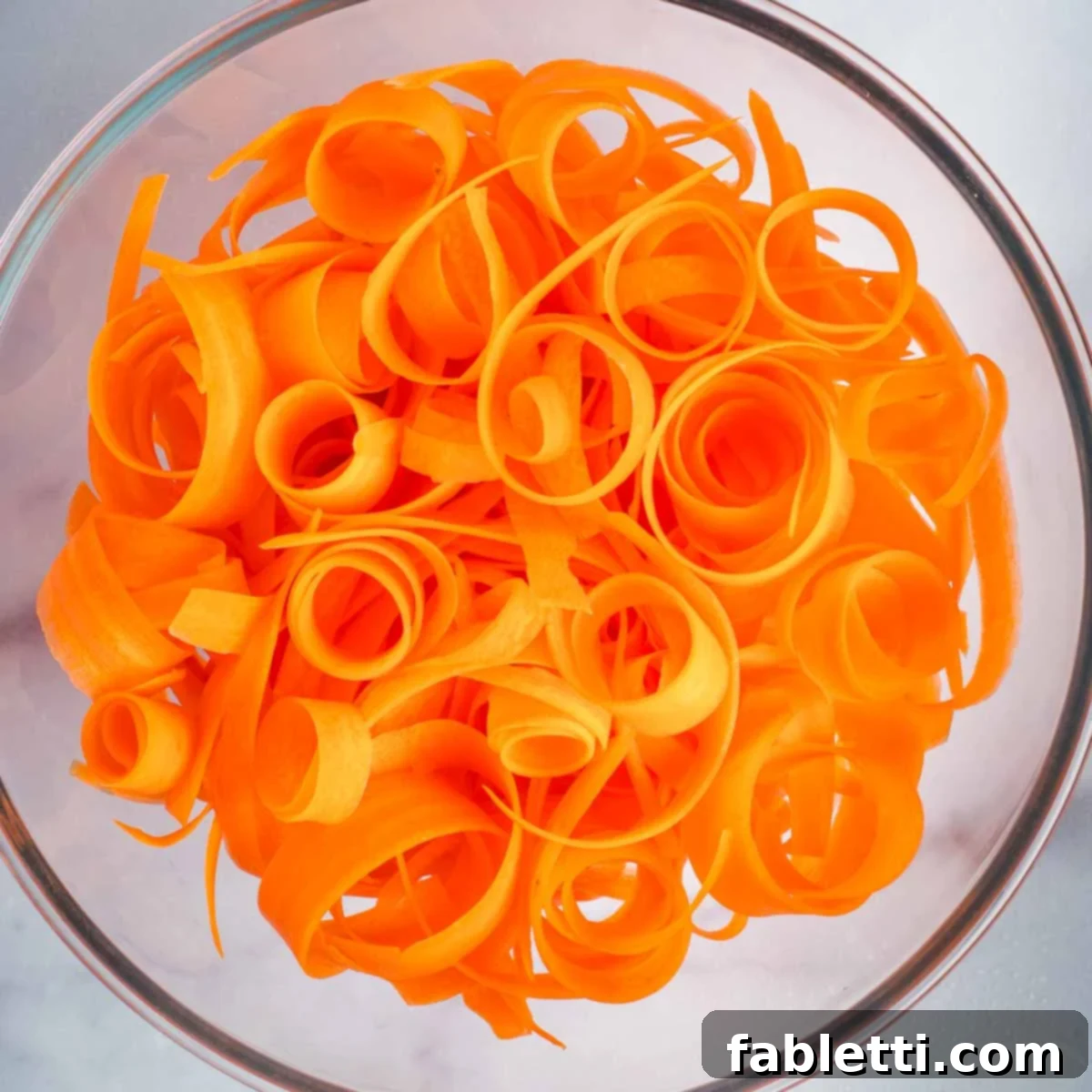 Sweet Heat Shaved Carrot Salad 6 Curly, vibrant orange carrot ribbons are neatly piled inside a clear glass mixing bowl, poised for the dressing and additional ingredients.