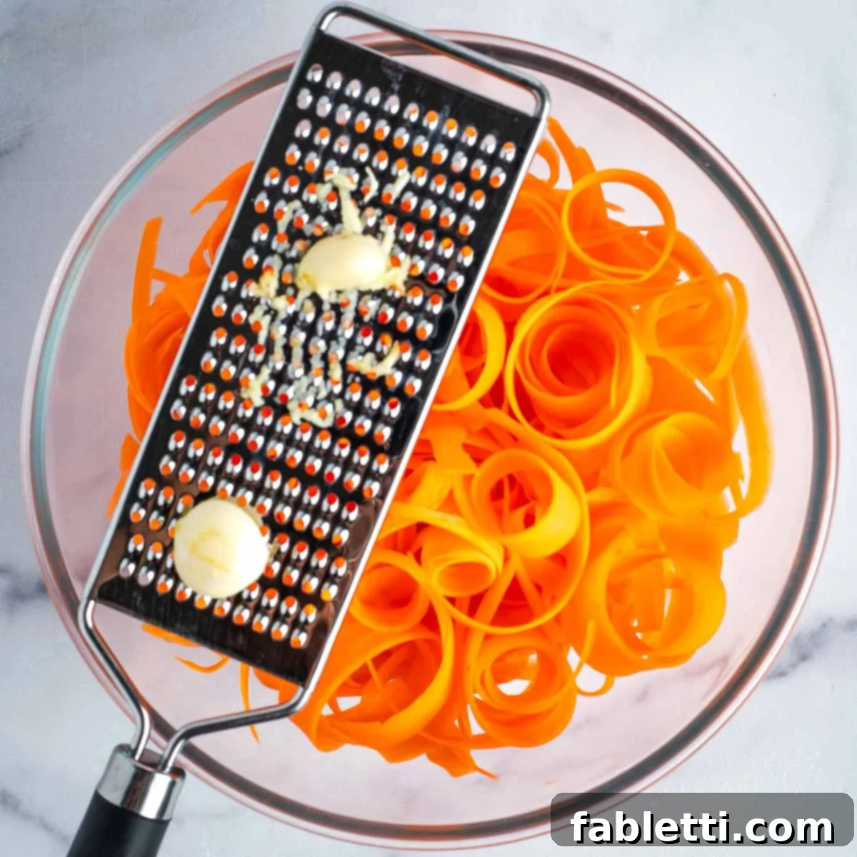 Sweet Heat Shaved Carrot Salad 7 A microplane grater is being used to finely grate fresh garlic cloves directly over a bowl filled with vibrant carrot ribbons, ensuring even distribution of flavor.