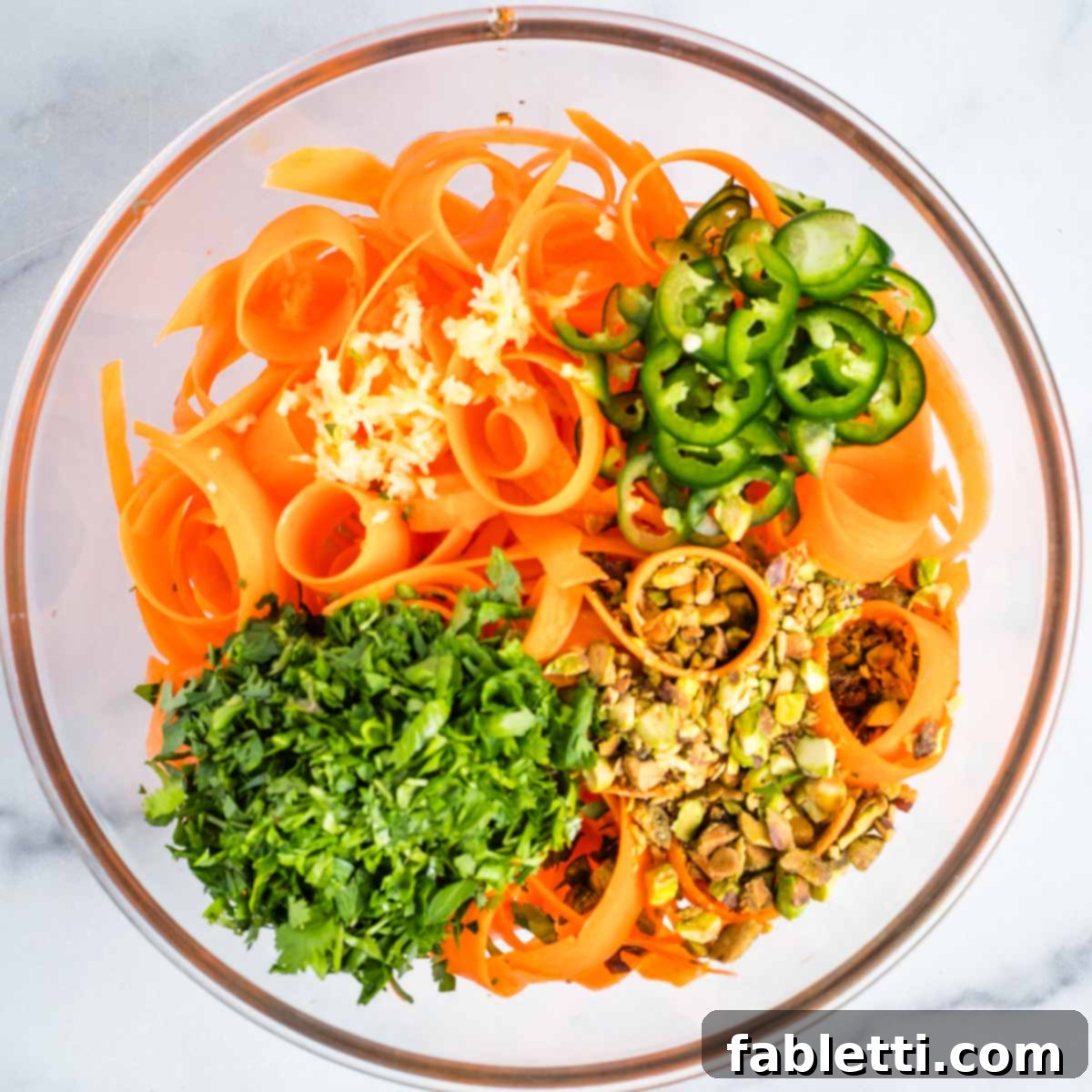 Sweet Heat Shaved Carrot Salad 8 A colorful bowl filled with carrot ribbons, generously topped with mounds of freshly grated garlic, finely chopped parsley, vibrant sliced jalapenos, and crunchy chopped pistachios, ready to be tossed.