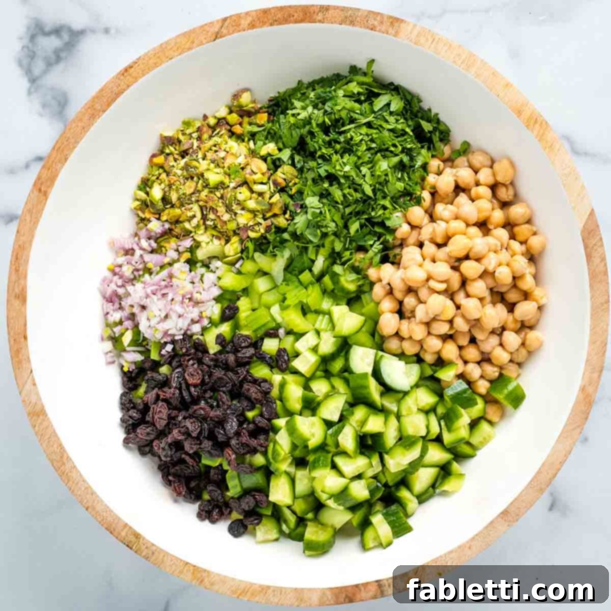 Crisp Celery & Chickpea Salad with a Sweet Raisin Twist 12 A vibrant salad bowl filled with chopped celery and cucumber, topped with neat piles of fresh parsley, rinsed chickpeas, sweet raisins, crunchy pistachios, and minced shallots, ready for the dressing.