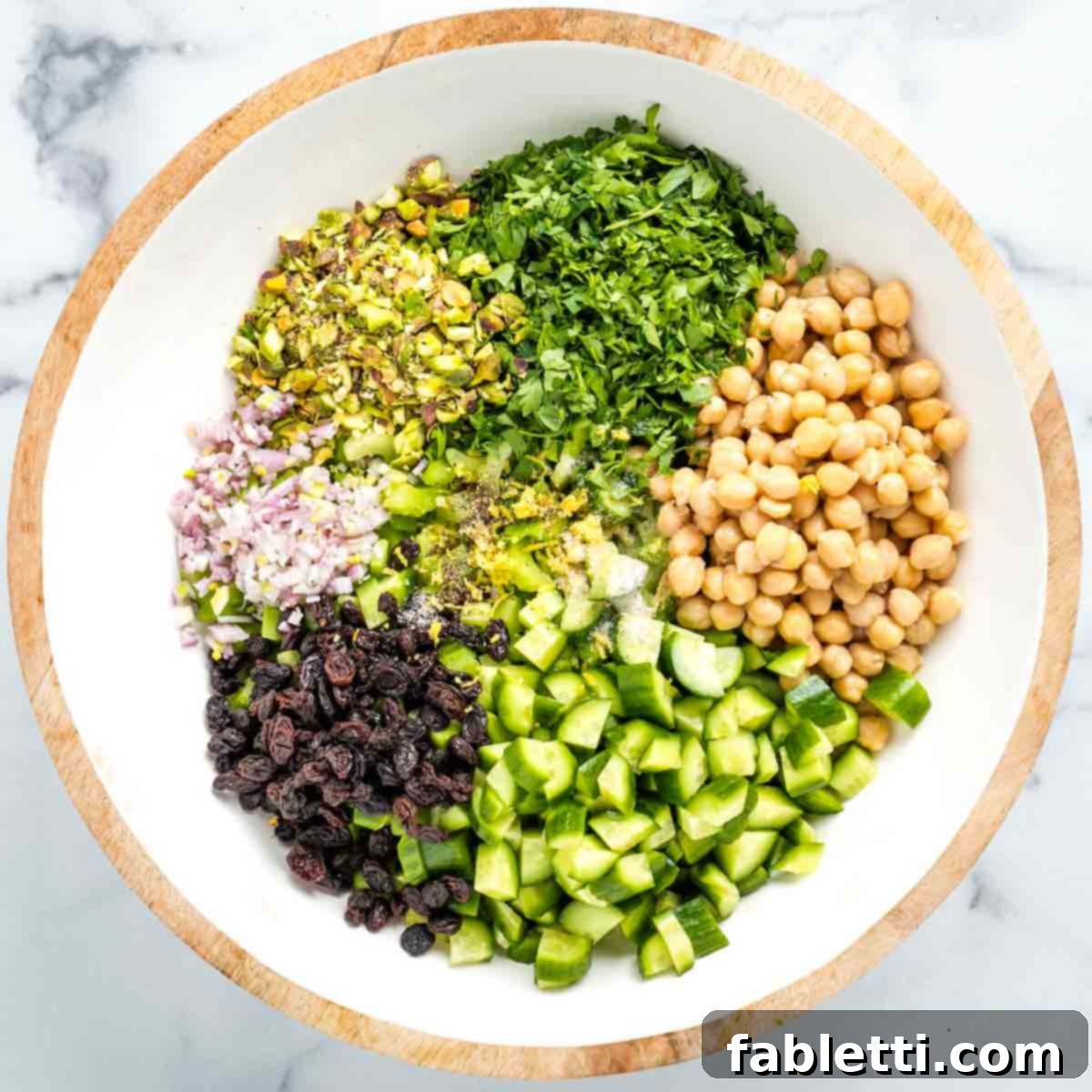 Crisp Celery & Chickpea Salad with a Sweet Raisin Twist 13 A close-up of a salad in a bowl, featuring chickpeas, cucumber, pistachios, fresh parsley, shallot, and raisins, being seasoned with fresh lemon zest, salt, and pepper, just before mixing.