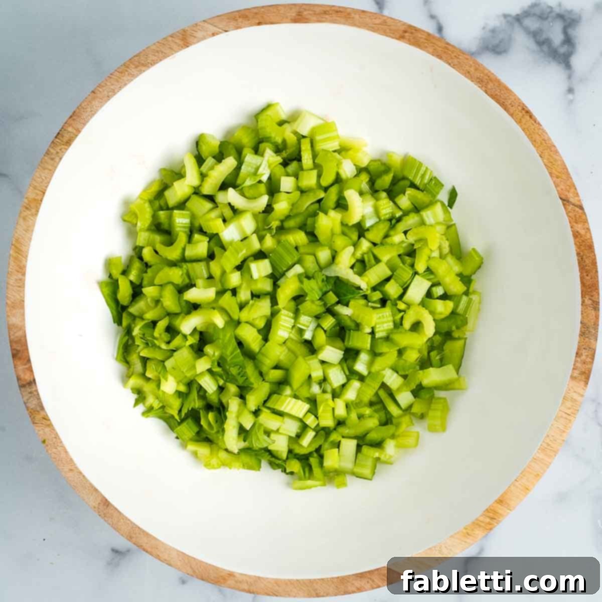 Crisp Celery & Chickpea Salad with a Sweet Raisin Twist 9 Finely diced celery pieces are gathered in an elegant white bowl with a wooden rim, awaiting other fresh salad ingredients.