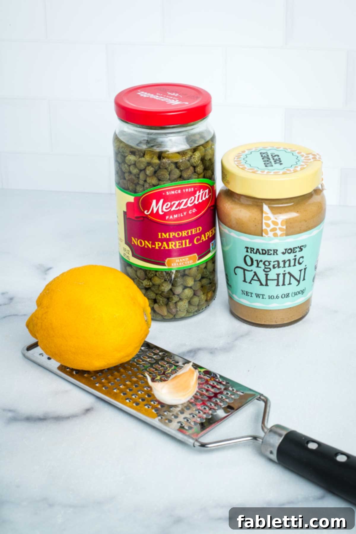 Jar of capers, jar of tahini, a lemon and a clove of garlic on a microplane grater.