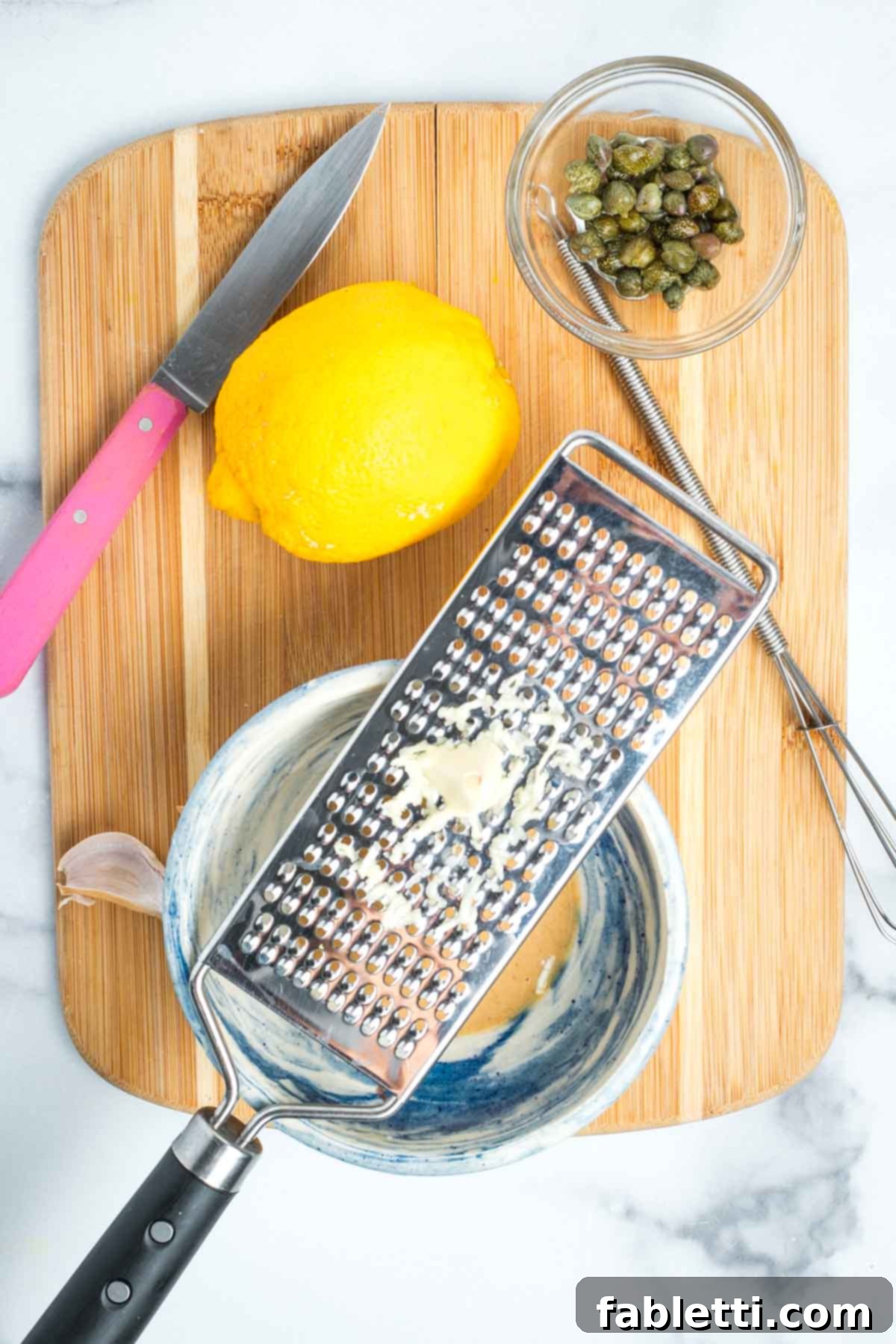 Zesting a clove of garlic over a bowl of tahini with lemon and caper on the cutting board.
