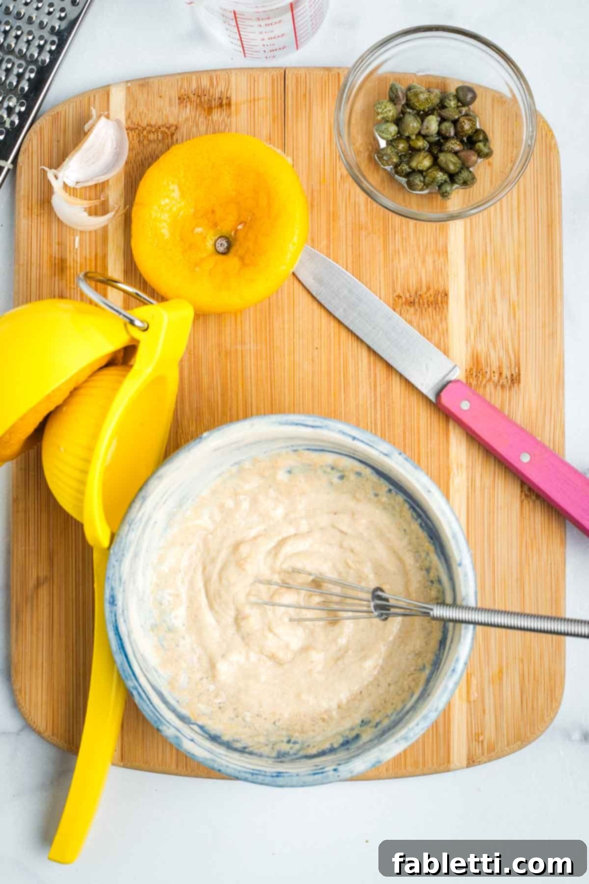White pasty mixture in a small bowl with lemons, garlic peel and capers strewn about.