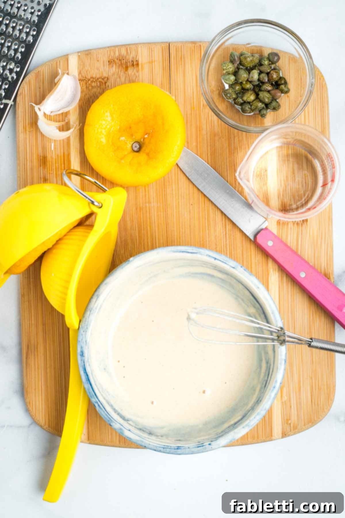 Messy work surface with lemons, capers, small dishes and a bowl of creamy sauce.