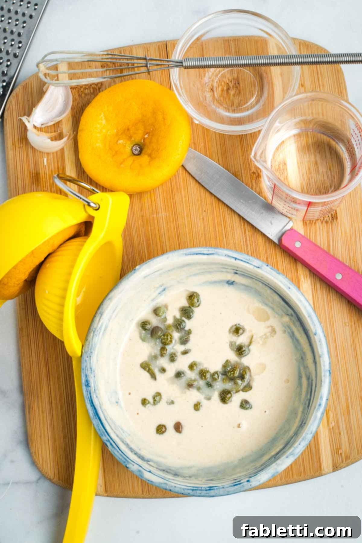 Small bowl with capers floating in a creamy sauce, ingredients strewn about on a cutting board.