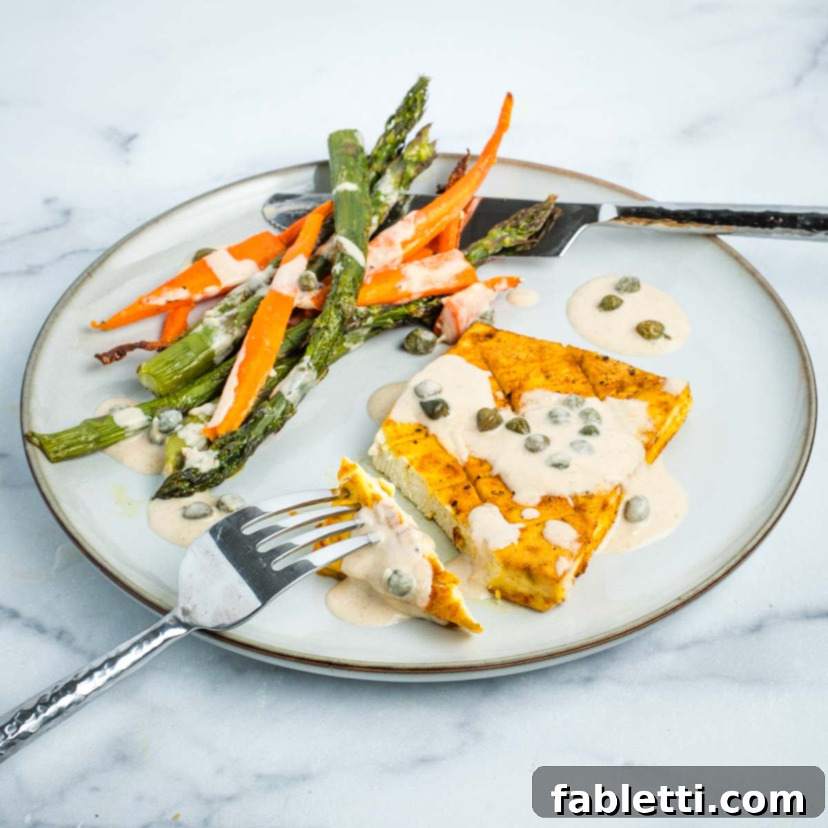 Cutting into a tofu steak drizzled with caper sauce on a plate with some carrots and asparagus.