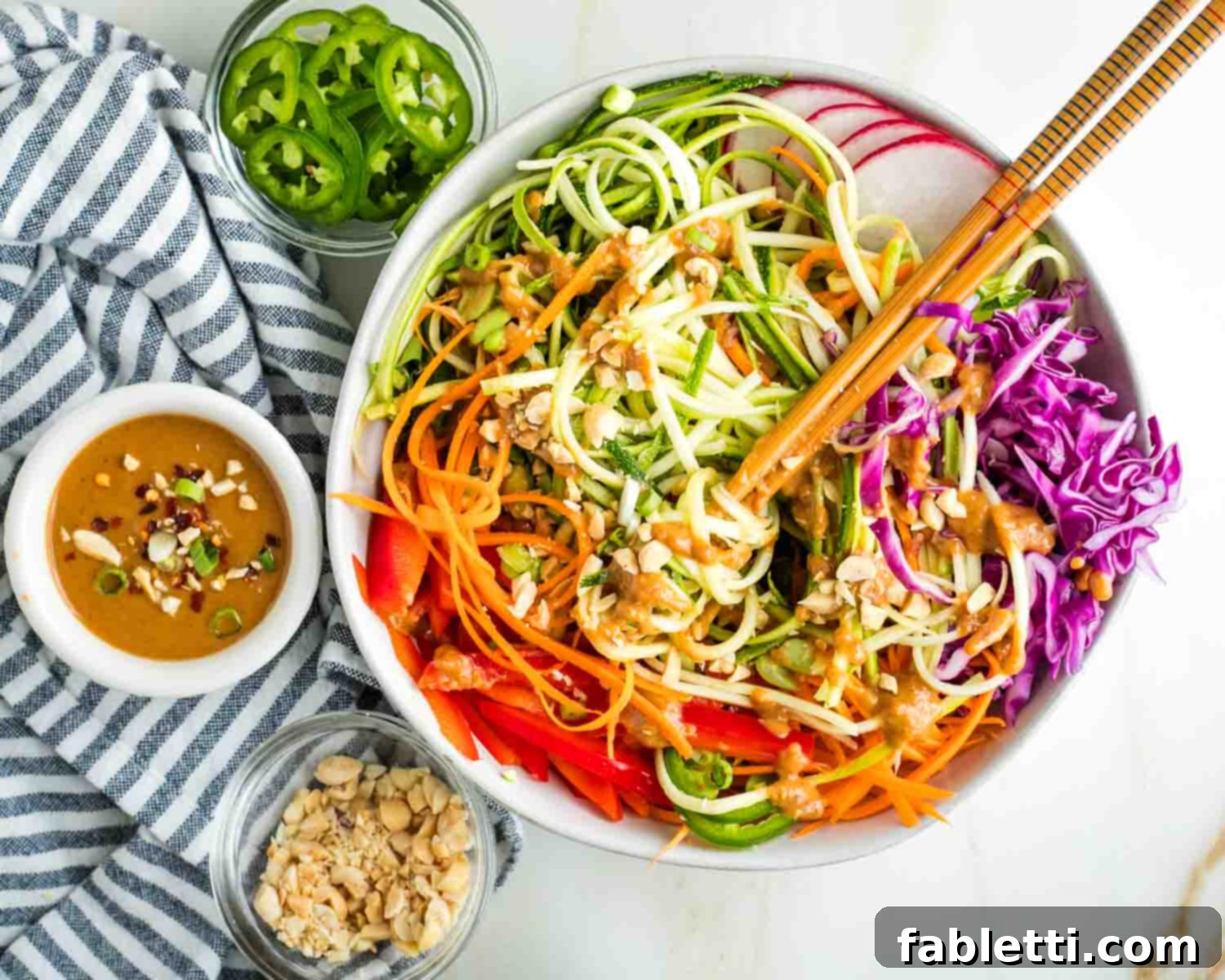 Fresh Spring Roll Salad Bowl with Creamy Peanut Sauce 19 Spring roll bowl with twirled zucchini pasta with chopsticks in a bowl full of vibrant veggies with peanut sauce, chopped peanuts and jalapenos.