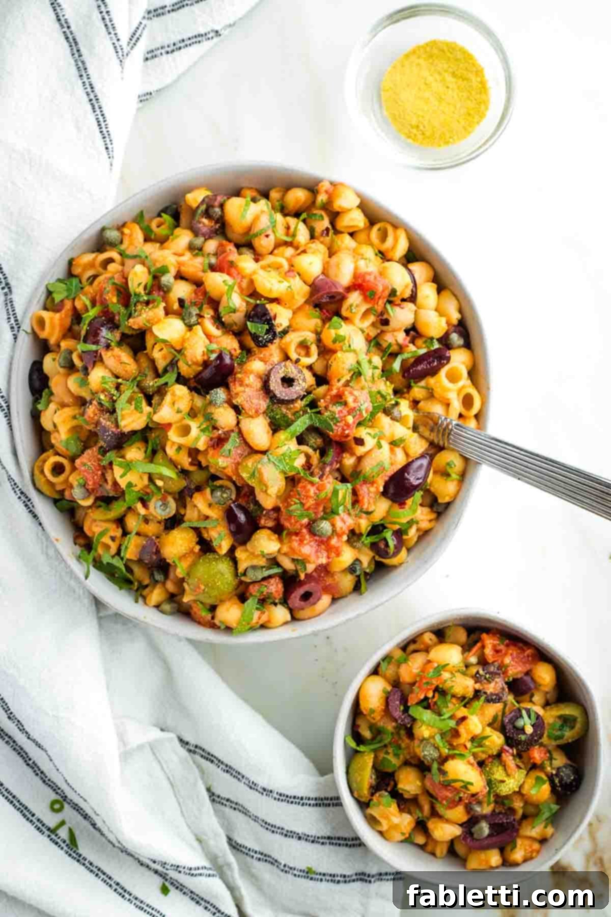 Zesty White Bean Puttanesca Pasta 2 One large and one small bowl of puttanesca pasta with white beans, olives and capers, garnished with fresh parsley.