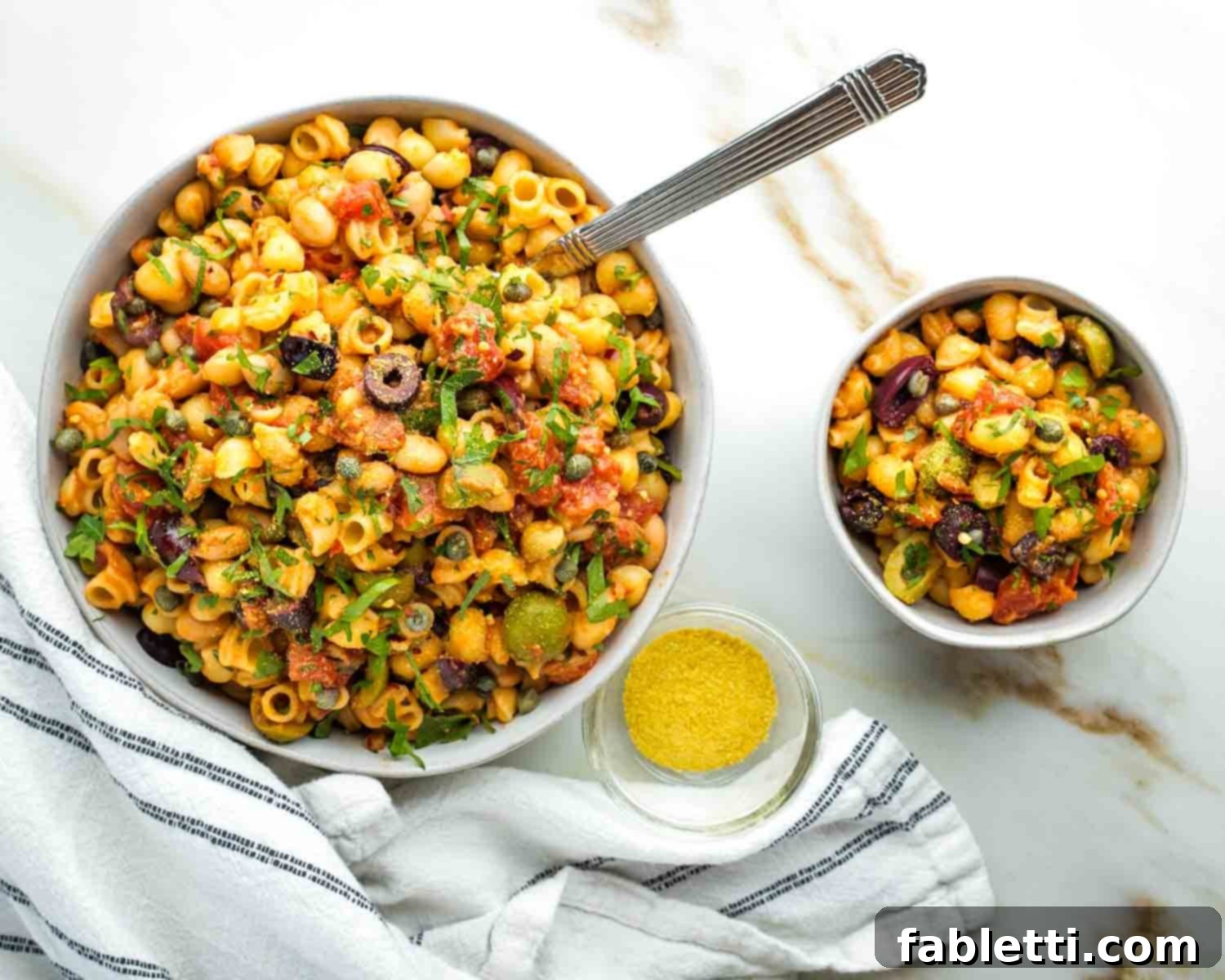 Zesty White Bean Puttanesca Pasta 12 Small and large bowls of vegan pasta puttanesca with white beans, olives, and capers, garnished with herbs.