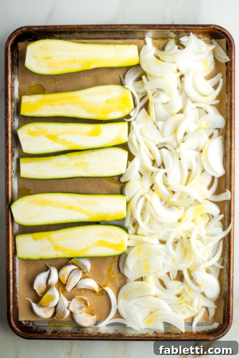 Caramelized Zucchini and Onion Creamy Dip 6 A baking tray filled with long halves of zucchini, thinly sliced onions, and whole garlic cloves, all generously drizzled with olive oil on parchment paper.