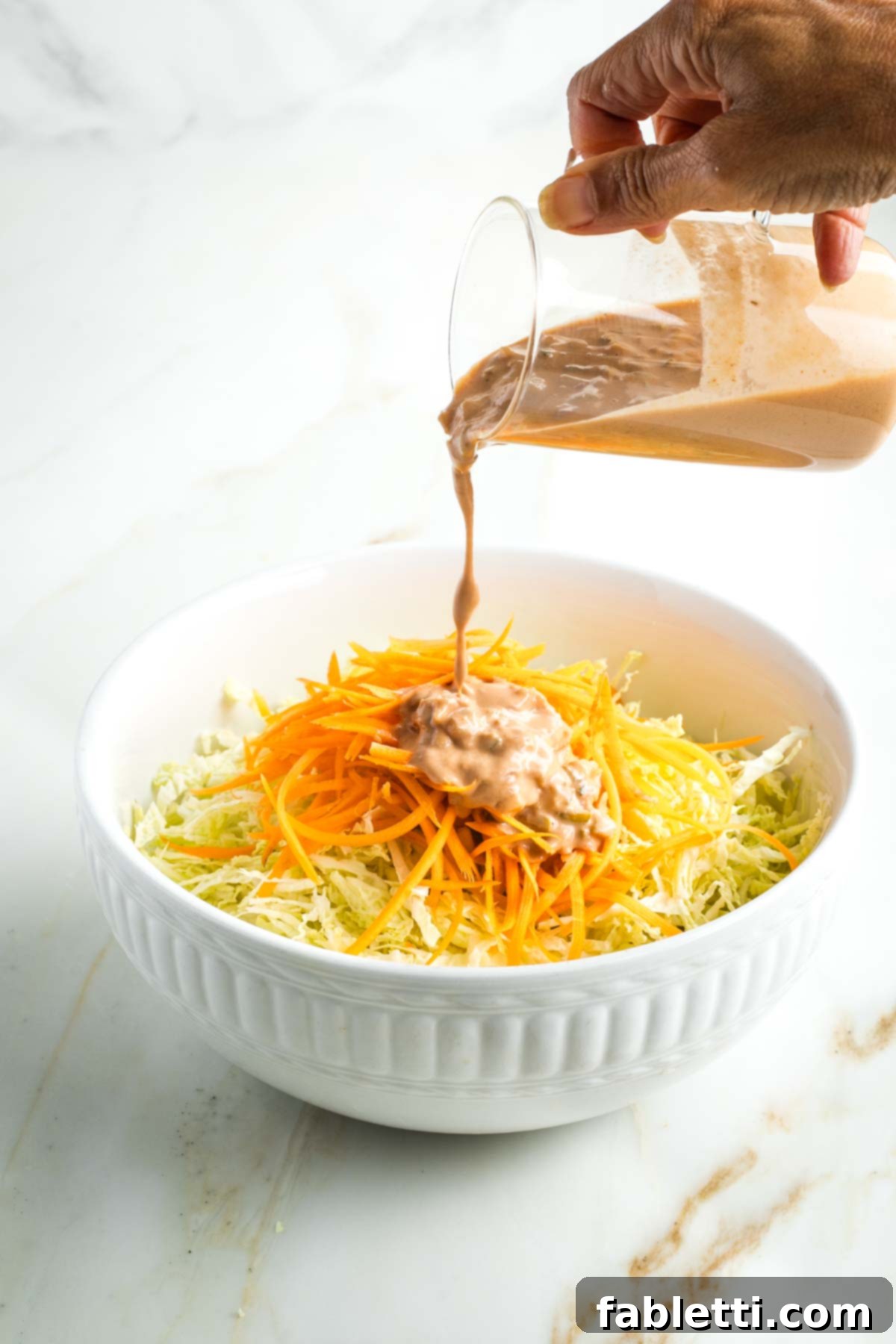 Pouring Russian dressing onto a bowl of shredded cabbage and carrots. 