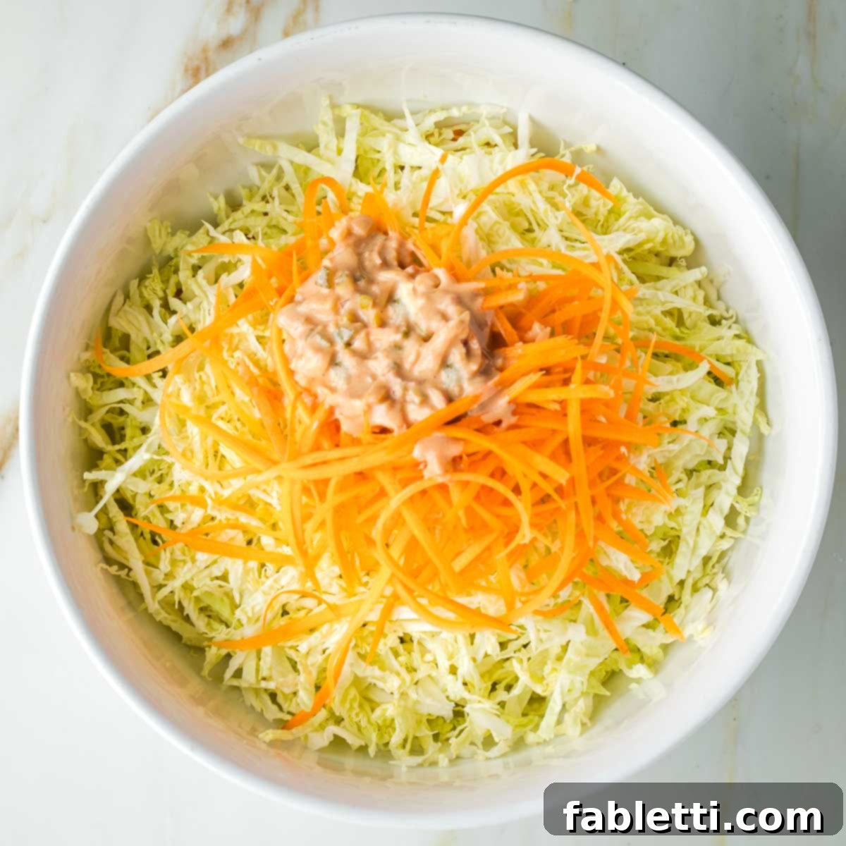 A pile of Russian dressing, chunky with pickles on top of a bowl of shredded cabbage and julienned carrots.