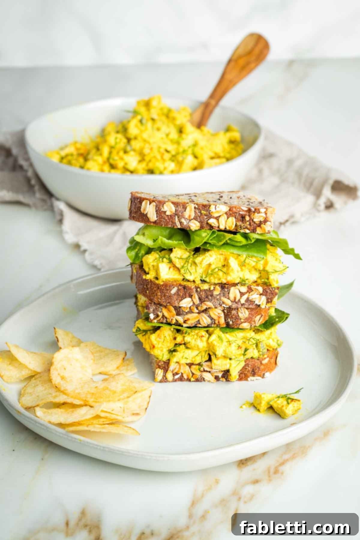 Zingy Vegan Tofu Egg Salad 2 Stacked vegan egg salad sandwich with chips on the plate and a bowl of the egg salad beyond.