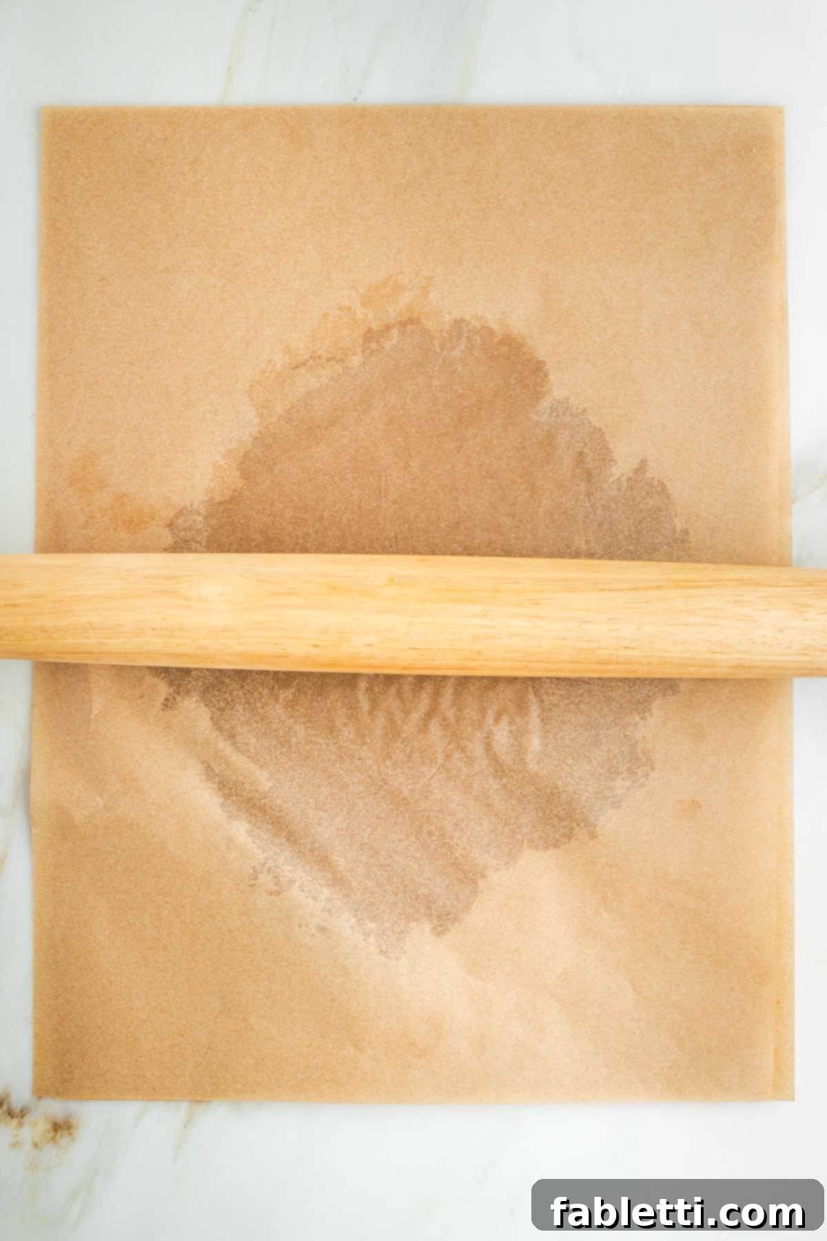 A rolling pin flattening one half of the pie dough into a large circular shape between two sheets of parchment paper.