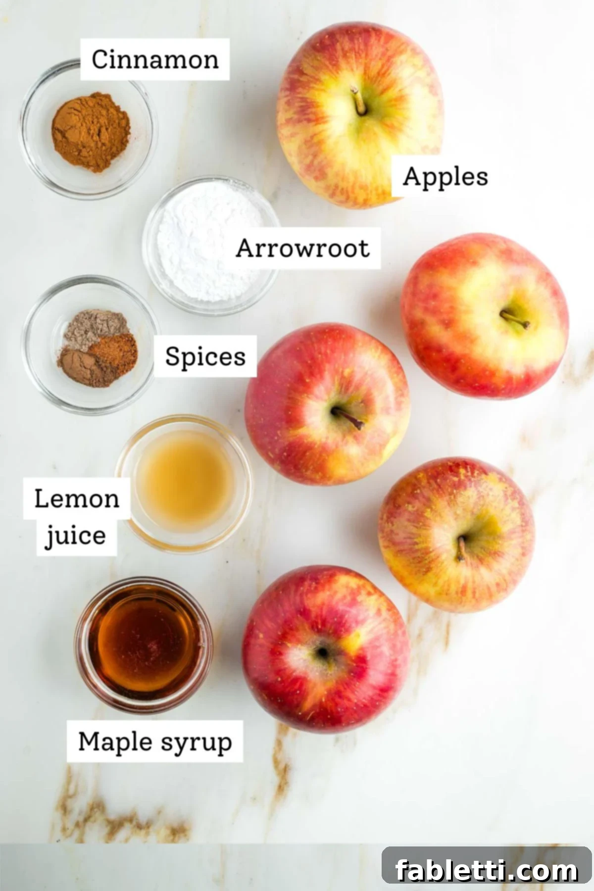 A selection of fresh Fuji apples, a bottle of maple syrup, lemon, and various spices including cinnamon, nutmeg, allspice, and cardamom, laid out for apple pie filling preparation.