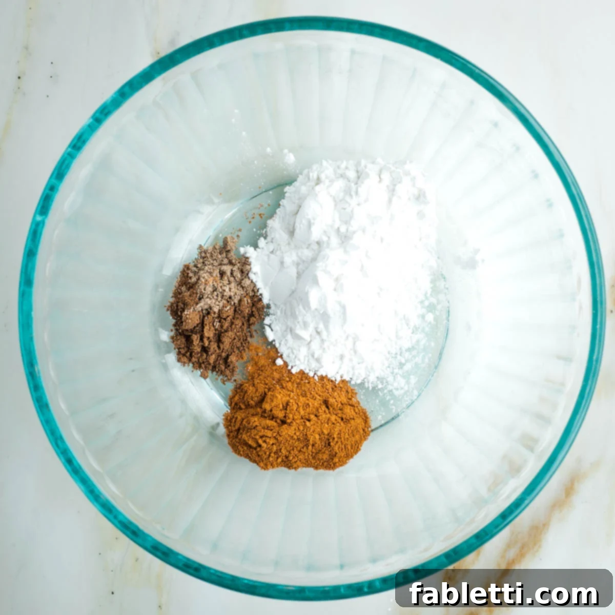 A glass bowl containing white arrowroot powder and various brown spices, ready to be combined.
