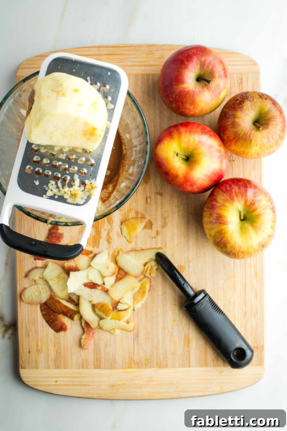 One apple being peeled and then grated directly into a glass bowl containing the liquid spice mixture, surrounded by whole apples and peels.