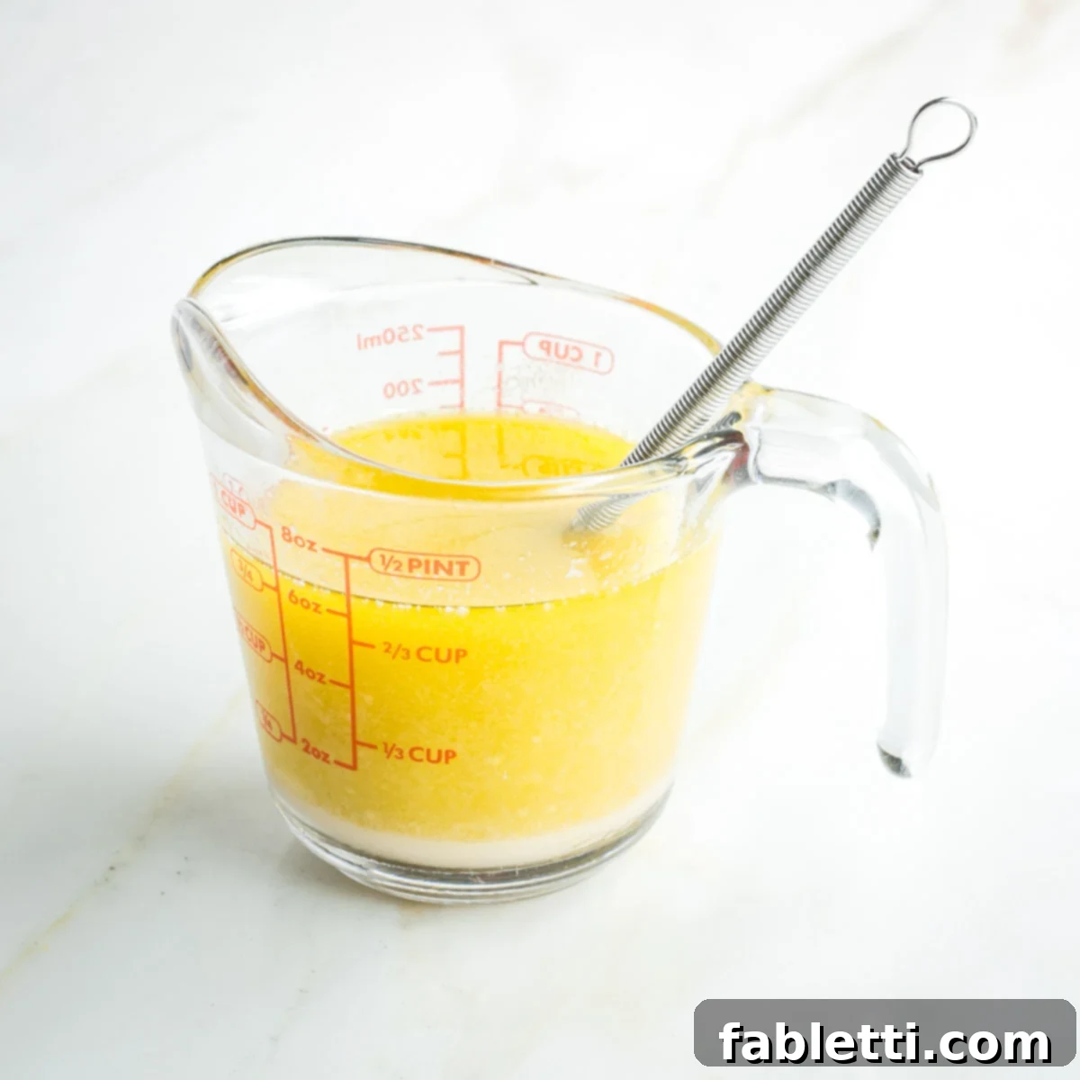 A small whisk mixing a yellow liquid inside a one-cup Pyrex measuring cup, creating an emulsified mixture.