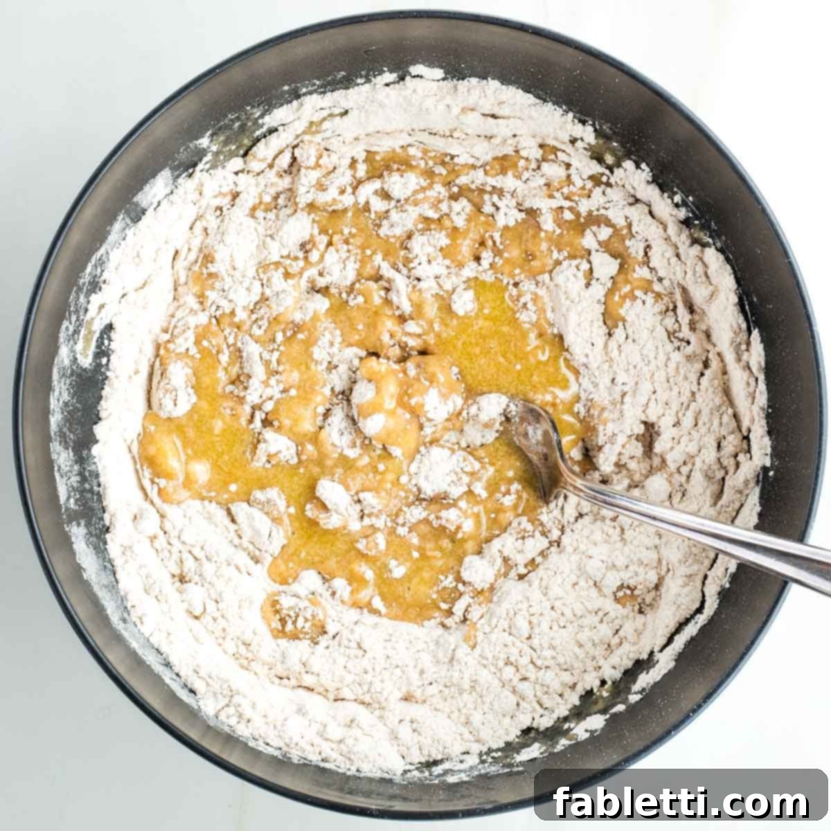 A fork mixing the yellow liquid into the flour mixture in a black bowl, forming a developing dough.